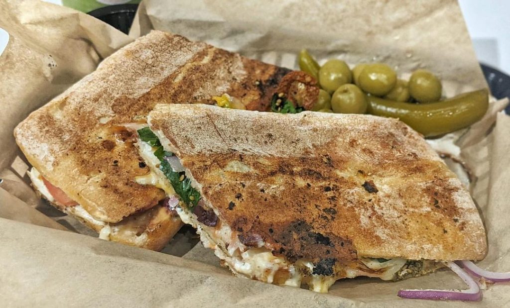a panini with a side of pickles and olives all in brown paper