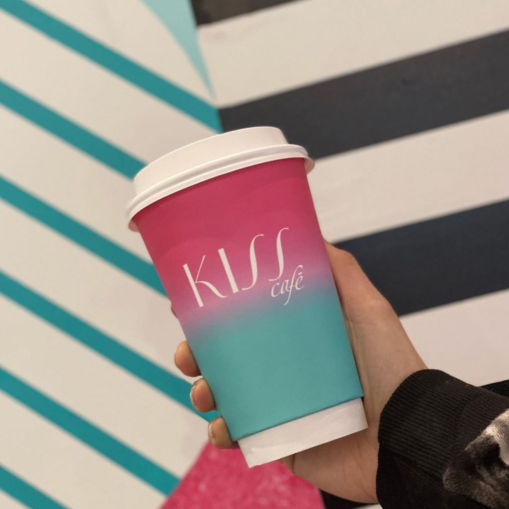 a hand holding a pink and blue coffee cup with the Kiss Cafe logo