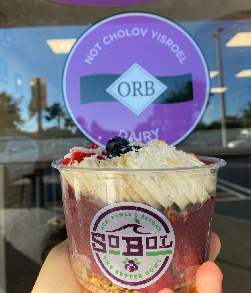 a hand holding an acai bowl in front of a purple sign