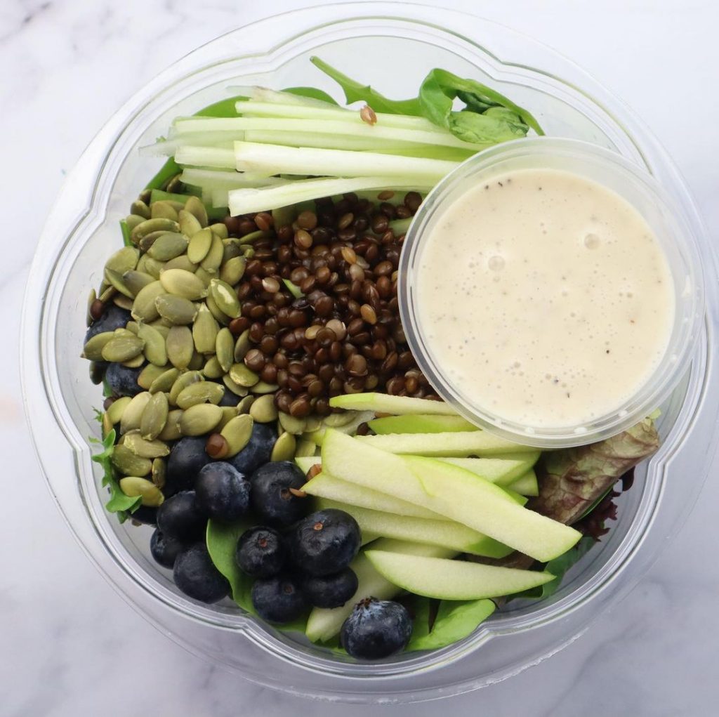 a salad with apple, blueberries, lentils, pumkin seeds and dressing in a cup